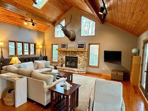 A cozy living room with a stone fireplace, a mounted deer head, and a large window overlooking a forest.