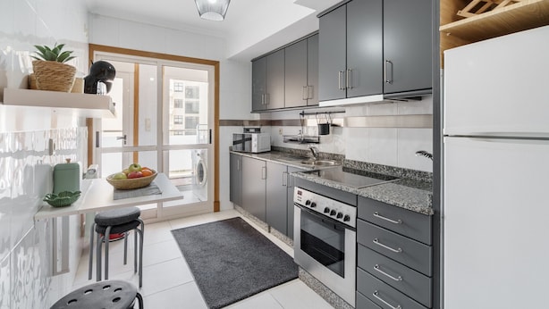 A modern kitchen with grey cabinets, a built-in oven, and a microwave. There is a window providing natural light and a view of the outside. - Sunny Beachside Escape by Minho s Guest (Povoa De Varzim)