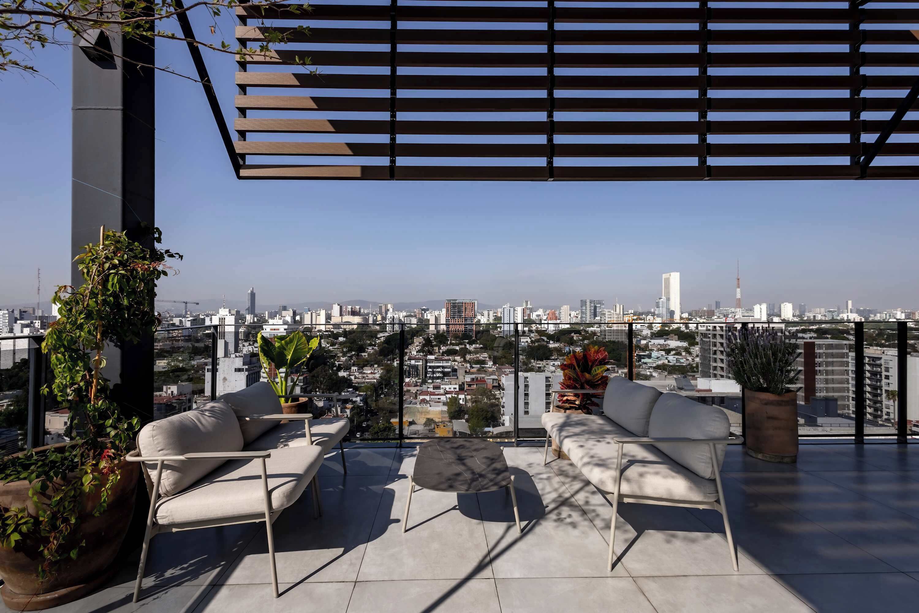 A rooftop terrace with a city view, furnished with two sofas and a small table, surrounded by potted plants.