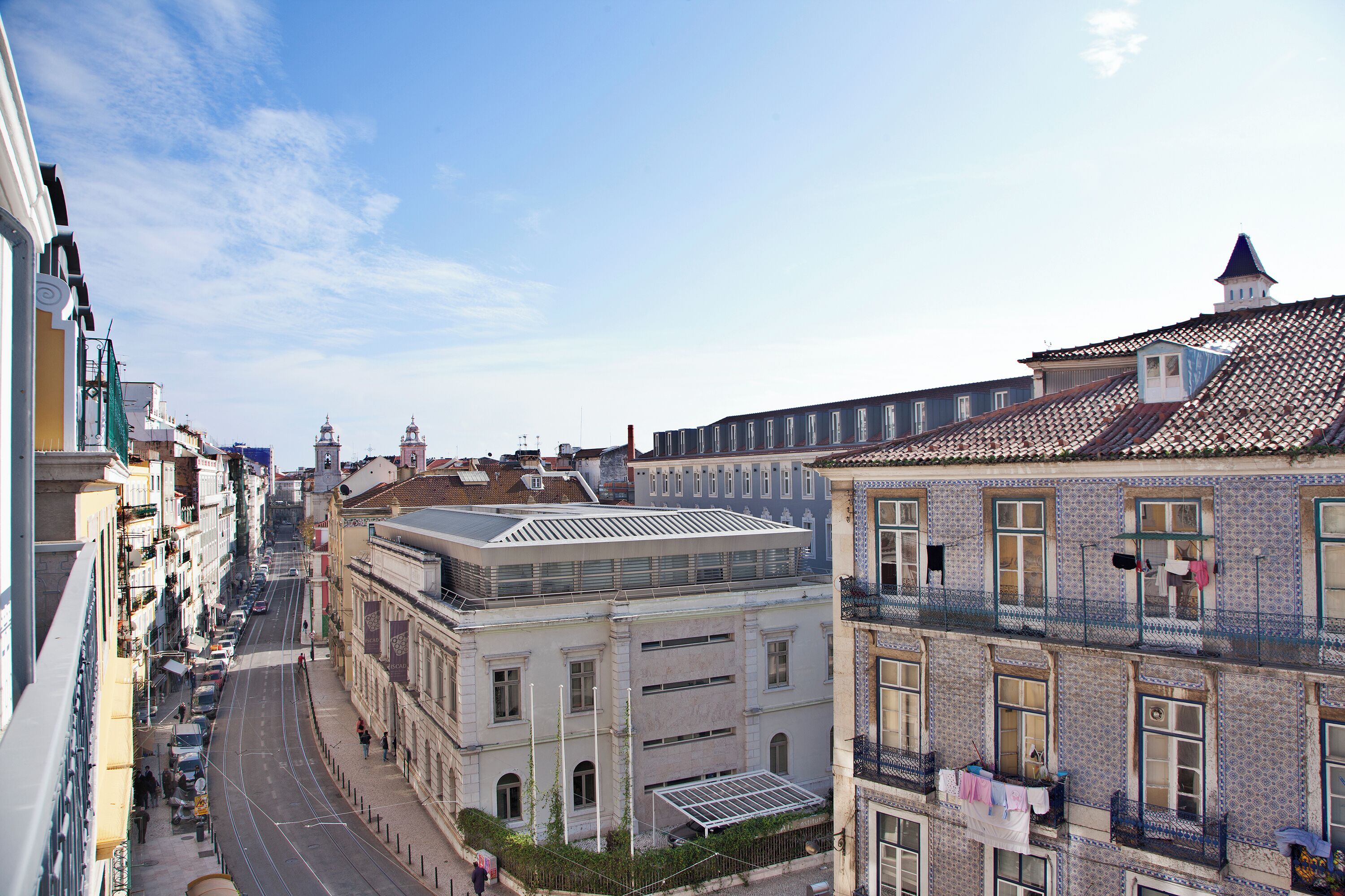 Stadtansicht mit Gebäuden, darunter ein markantes Gebäude mit flachem Dach und einem Balkon, an dem Wäsche aufgehängt ist.