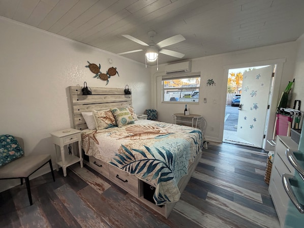 Un dormitorio con cabecera de madera, cama con colcha estampada con motivos de hojas, ventilador de techo y ventana con vistas al exterior.