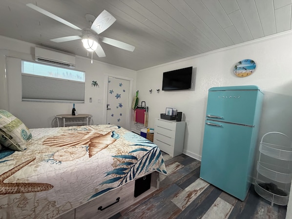 Ein Schlafzimmer mit Bett, Deckenventilator, Fernseher, Kühlschrank und einem Fenster mit Jalousien.