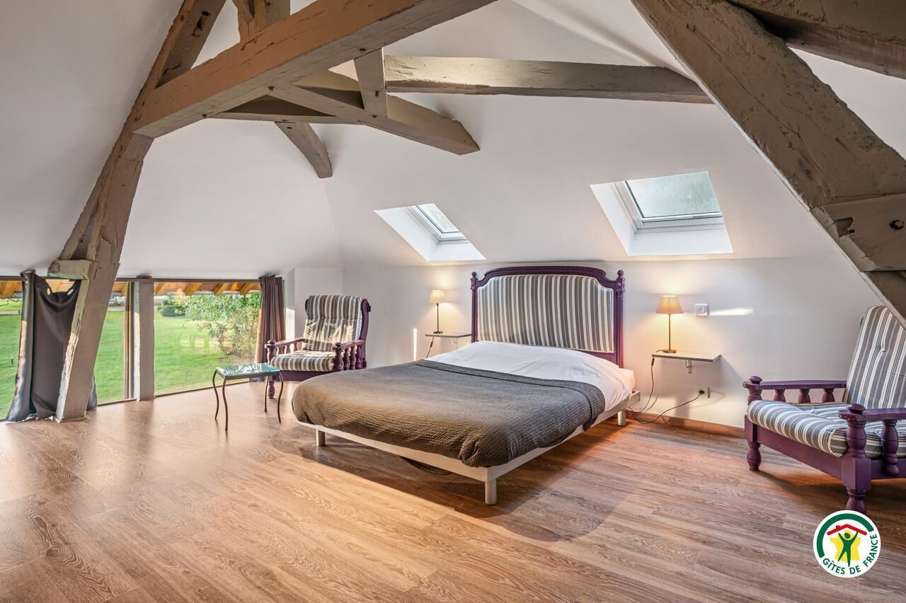 Ein Schlafzimmer mit Holzbalkendecke, einem Bett, zwei Stühlen und Blick auf einen Garten durch große Fenster.