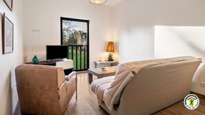 A modern living room with a sofa, a television, a lamp, and a view of a building through a window. - Gîtes de France® - Le Gîte du Château (BERTREVILLE SAINT OUEN)
