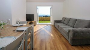 Ein modernes Wohnzimmer mit einem Holzesstisch, gedeckt für zwei Personen, einem grauen Sofa und einem Fernseher auf einem Holzschrank.