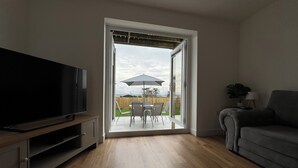 Eine offene Tür führt auf eine Terrasse mit Tisch und Stühlen, links ein Fernseher und rechts eine Couch.