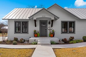 A single-story house with a grey exterior, numbered 205, featuring a front entrance with a grey door, two windows on either side, and a small porch area with potted plants. - Miles Manor - Pool, 2 Hot Tubs & Walk to Main (Fredericksburg)