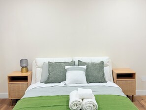 Un letto ben rifatto con una coperta verde, due cuscini e asciugamani arrotolati su un comodino in legno.