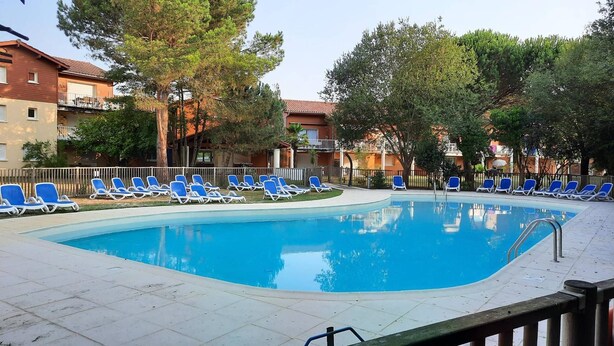 A swimming pool with blue lounge chairs arranged around it, surrounded by trees and buildings. - AMARINE Residence – 2-Bedroom Apartment, Terrace, Heated Pool  et  WiFi (CAPBRETON)
