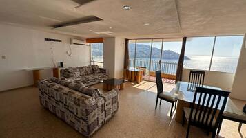 A spacious living room with a sofa, a dining table, and chairs. There is a large window offering a view of the sea and hills.