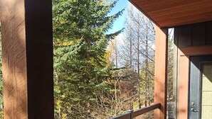 A wooden balcony with a metal railing, a concrete floor, and a view of trees and a building in the background. - Stunning 2-BR Suite with Hot Tub & Mountain Views (Canmore)