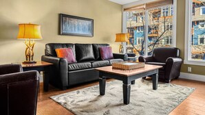 A living room with a black leather sofa, two side tables with lamps, a coffee table, and a large window with a view of buildings. - Elegant Mountain Retreat | Pool and Hot Tub (Canmore)