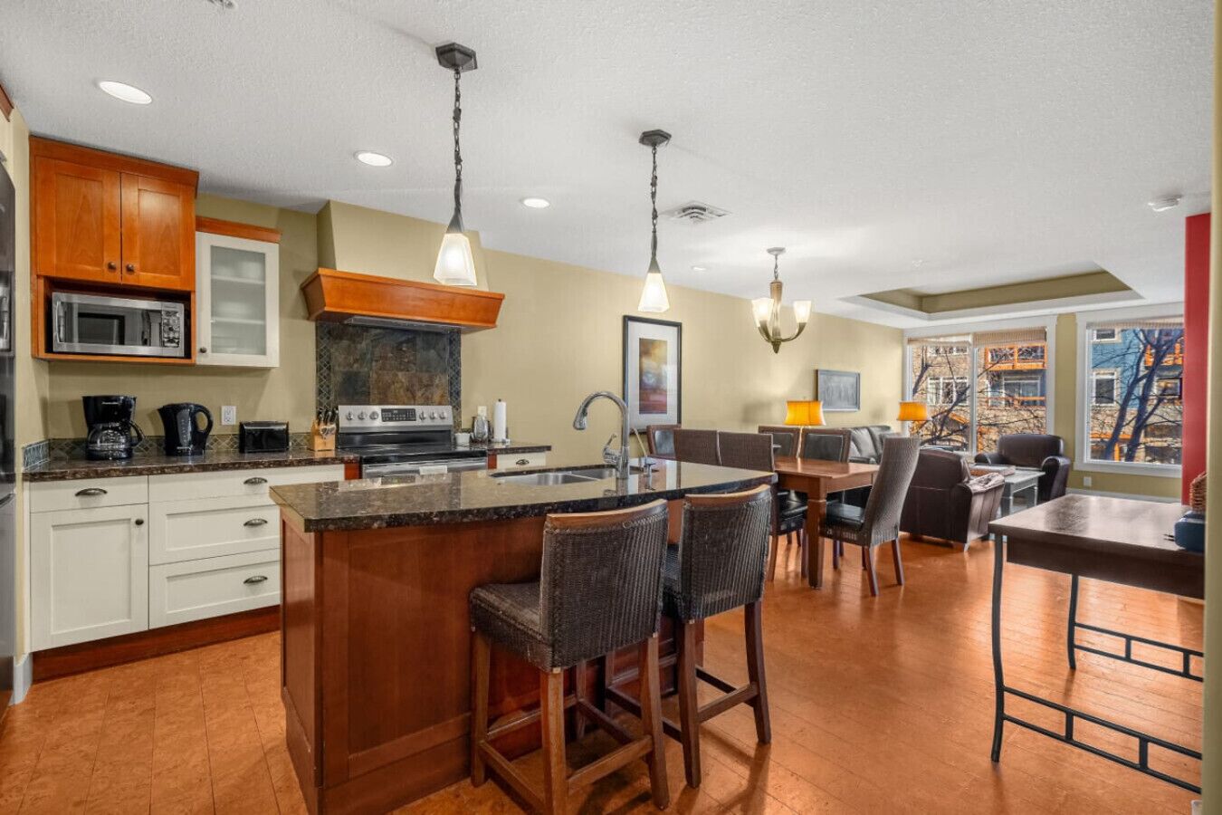 A modern kitchen with a central island, bar stools, a dining area with a table and chairs, and a view of the outdoors through large windows.