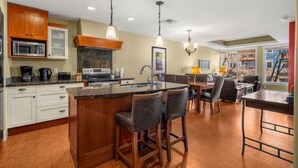 A modern kitchen with a central island, bar stools, a dining area with a table and chairs, and a view of the outdoors through large windows. - Elegant Mountain Retreat | Pool and Hot Tub (Canmore)