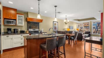 A modern kitchen with a central island, bar stools, a dining area with a table and chairs, and a view of the outdoors through large windows.