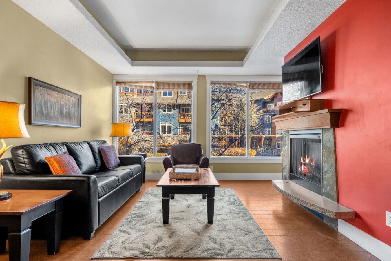 A living room with a fireplace, a sofa, a coffee table, and a view of buildings through large windows.