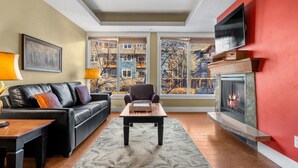 A living room with a fireplace, a sofa, a coffee table, and a view of buildings through large windows. - Elegant Mountain Retreat | Pool and Hot Tub (Canmore)
