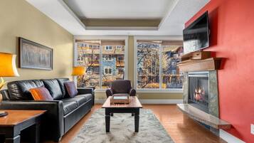 A living room with a fireplace, a sofa, a coffee table, and a view of buildings through large windows.