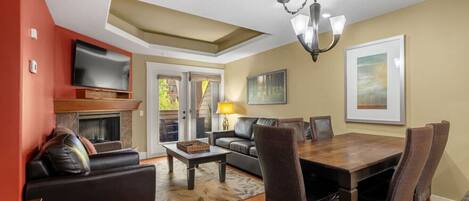 Een moderne woonkamer met een open haard, een televisie, een eethoek met een tafel en stoelen, en een groot raam met uitzicht op de omgeving.