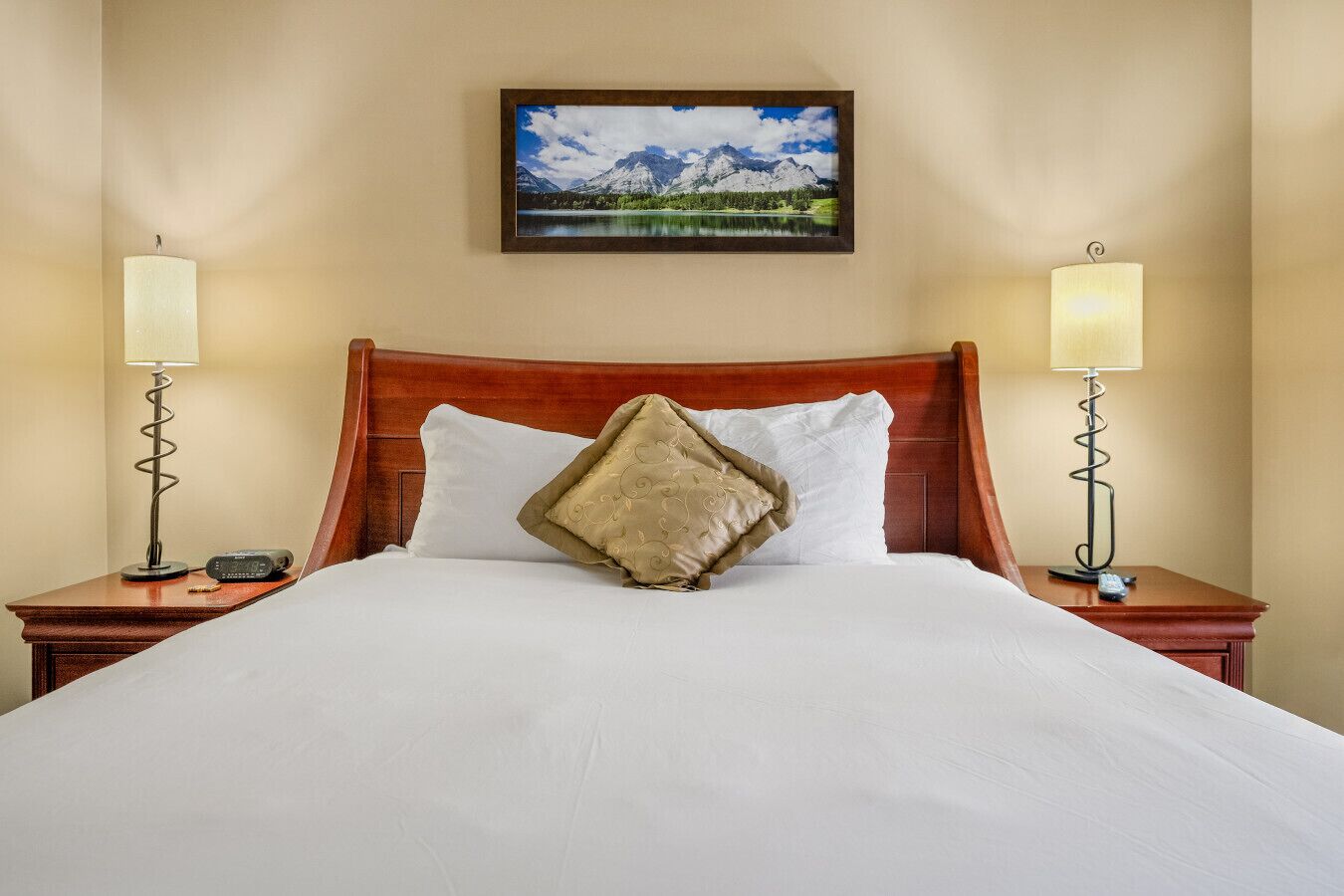 A neatly made bed with white linens, a wooden headboard, and a single decorative pillow. Two bedside lamps with spiral bases are on either side of the bed.