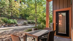 A covered patio with a table and chairs, surrounded by trees and a stone wall. - Modern Condo Retreat 2BR+Sofa Bed | Patio & Trails (Canmore)