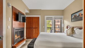 Ein Schlafzimmer mit Kamin, einem Bett, einem an der Wand befestigten Fernseher und einer Balkontür.