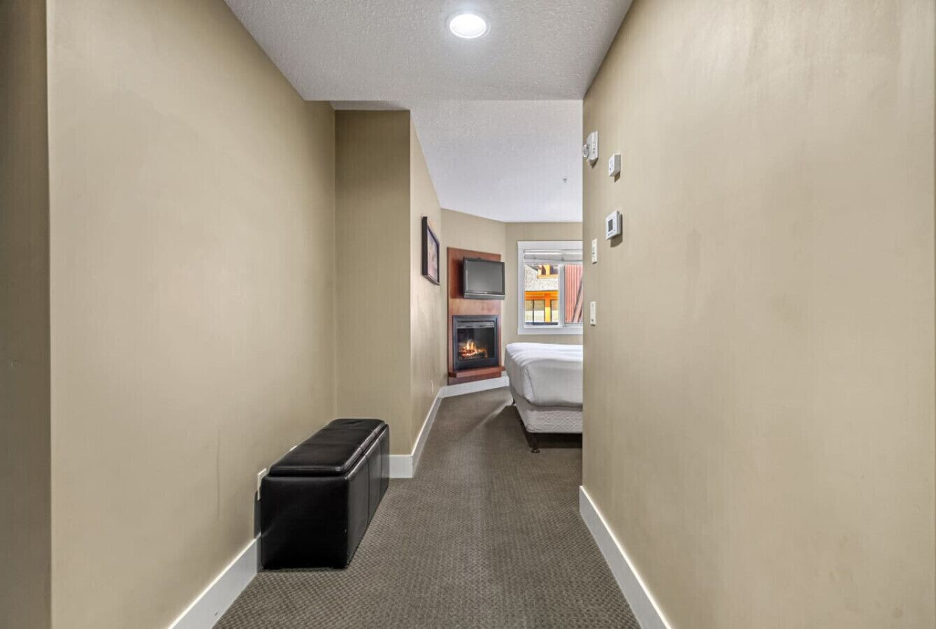 A narrow hallway with beige walls, a carpeted floor, a black storage box, a fireplace, and a bed with a white headboard.