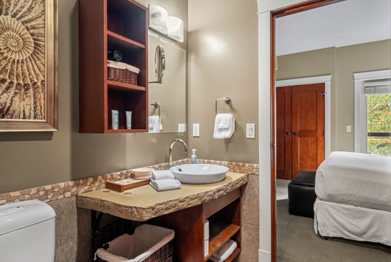 A bathroom with a sink, mirror, and shelves with toiletries. There is a door leading to a bedroom with a bed covered in white sheets.