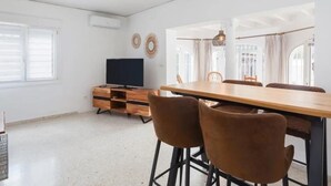 Un vaste espace de vie comprenant une table à manger, deux chaises marron, un téléviseur posé sur un meuble en bois et une grande fenêtre.