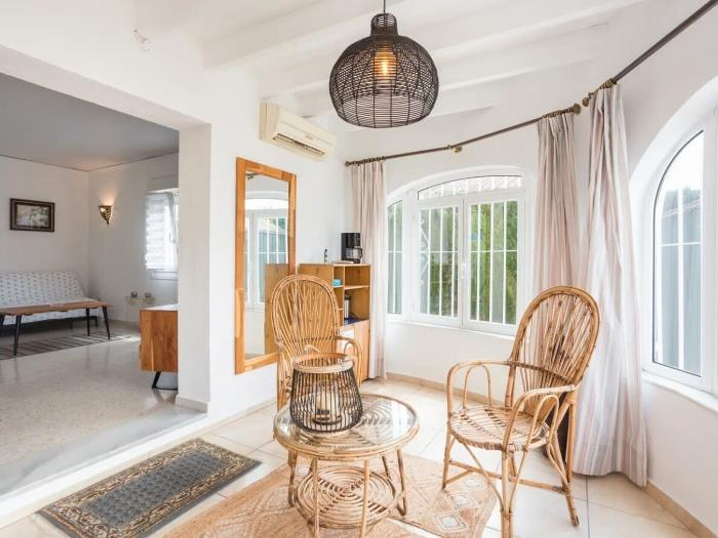 A spacious room with a large window, a round table with chairs, a bookshelf, and a mirror on the wall.