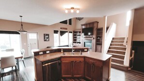A modern kitchen with a built-in dishwasher, a large island with a sink, wooden cabinets, and a staircase leading to another level. - Lake Community Retreat - Family Parks (Calgary)