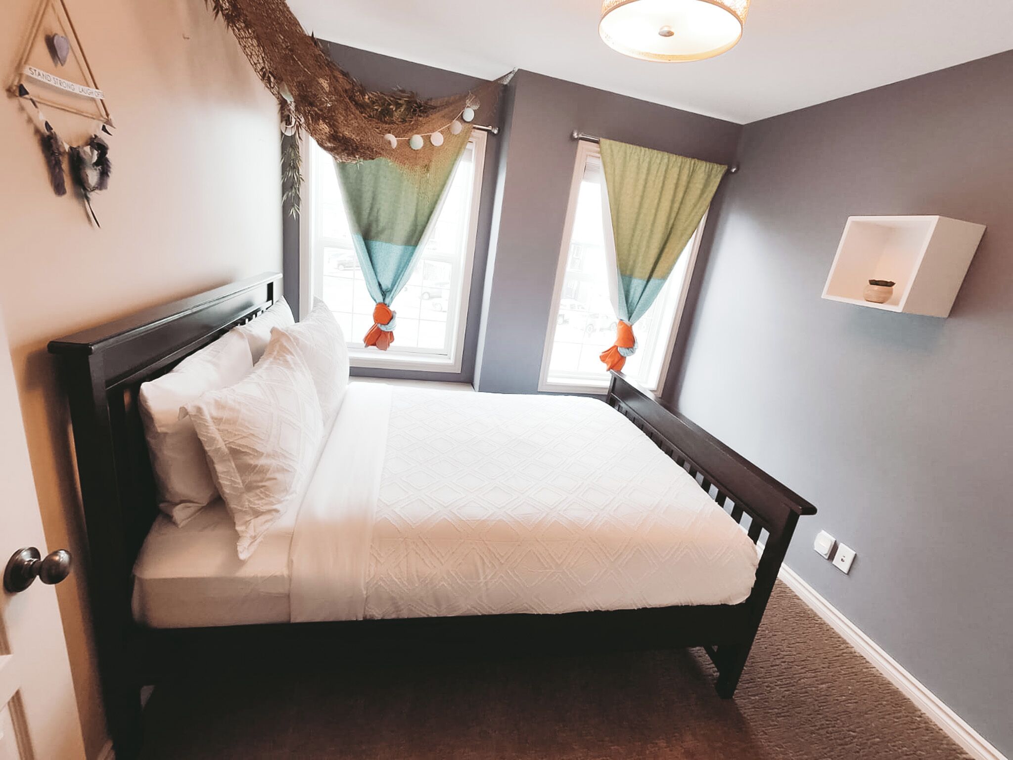A bedroom with a bed, two windows with curtains, a wall-mounted shelf, and a door.