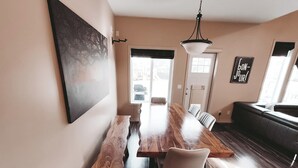 A modern dining area with a long wooden table, chairs, and a bench. There is a black sofa and a wall-mounted light fixture. - Lake Community Retreat - Family Parks (Calgary)