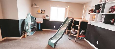 A children's playroom with a slide, shelves with toys, and a window.