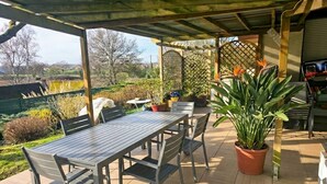 Um pátio externo com mesa e cadeiras, rodeado por plantas em vasos e vista para o campo.