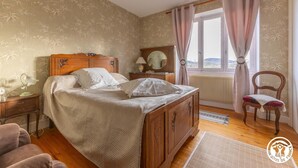Ein Schlafzimmer mit einem hölzernen Bett, einem Nachttisch, einem Sessel, einem Fenster mit Vorhängen und einer Heizung.