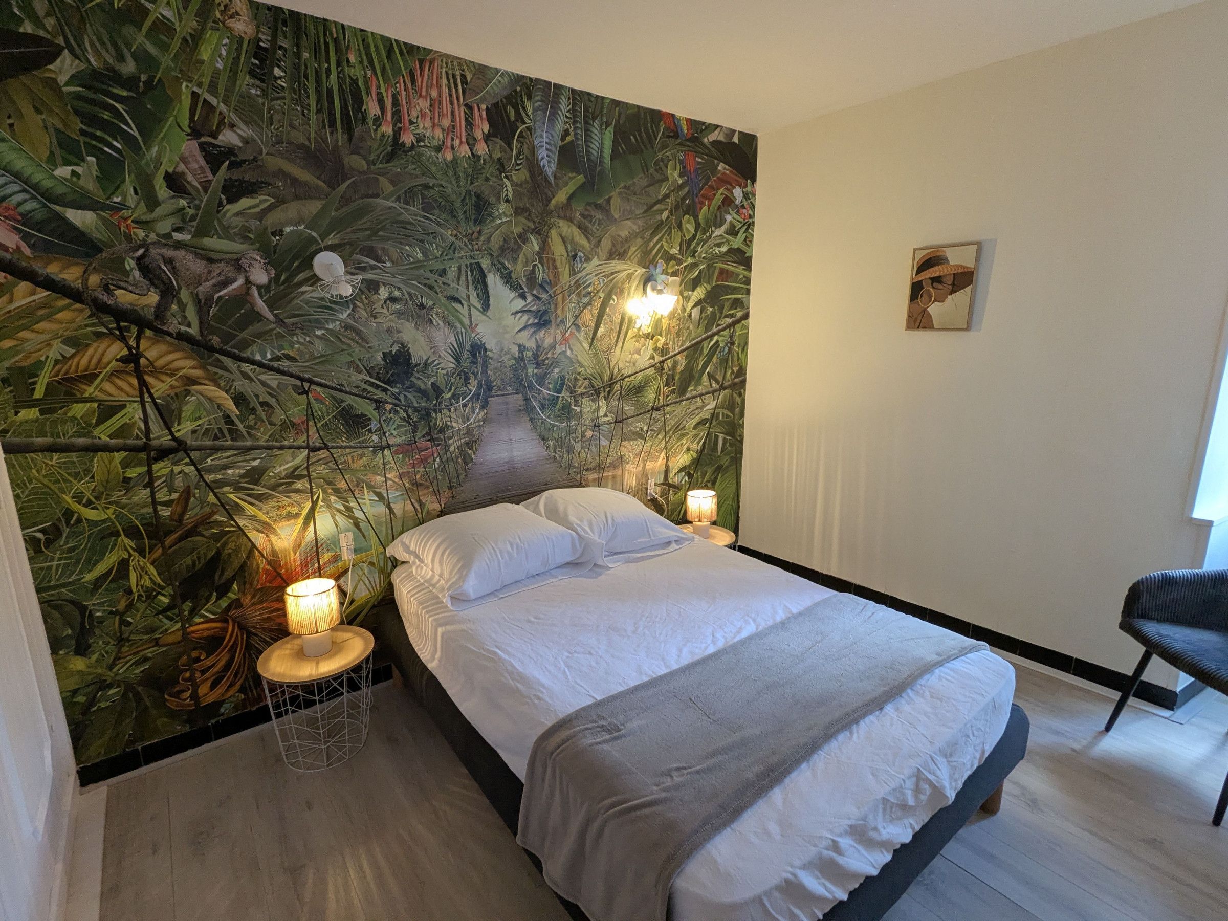 Et soveværelse med et vægmaleri af en jungle, en seng med hvide sengetøj, to sengeborde med lamper og en stol.