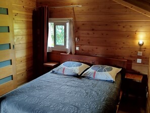 A wooden cabin room with a bed, a window with curtains, and a wall-mounted light.