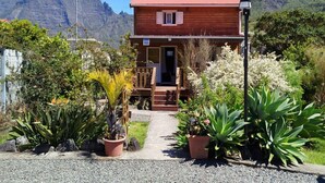 A wooden house with a red roof, surrounded by a garden with various plants and a mountain in the background.