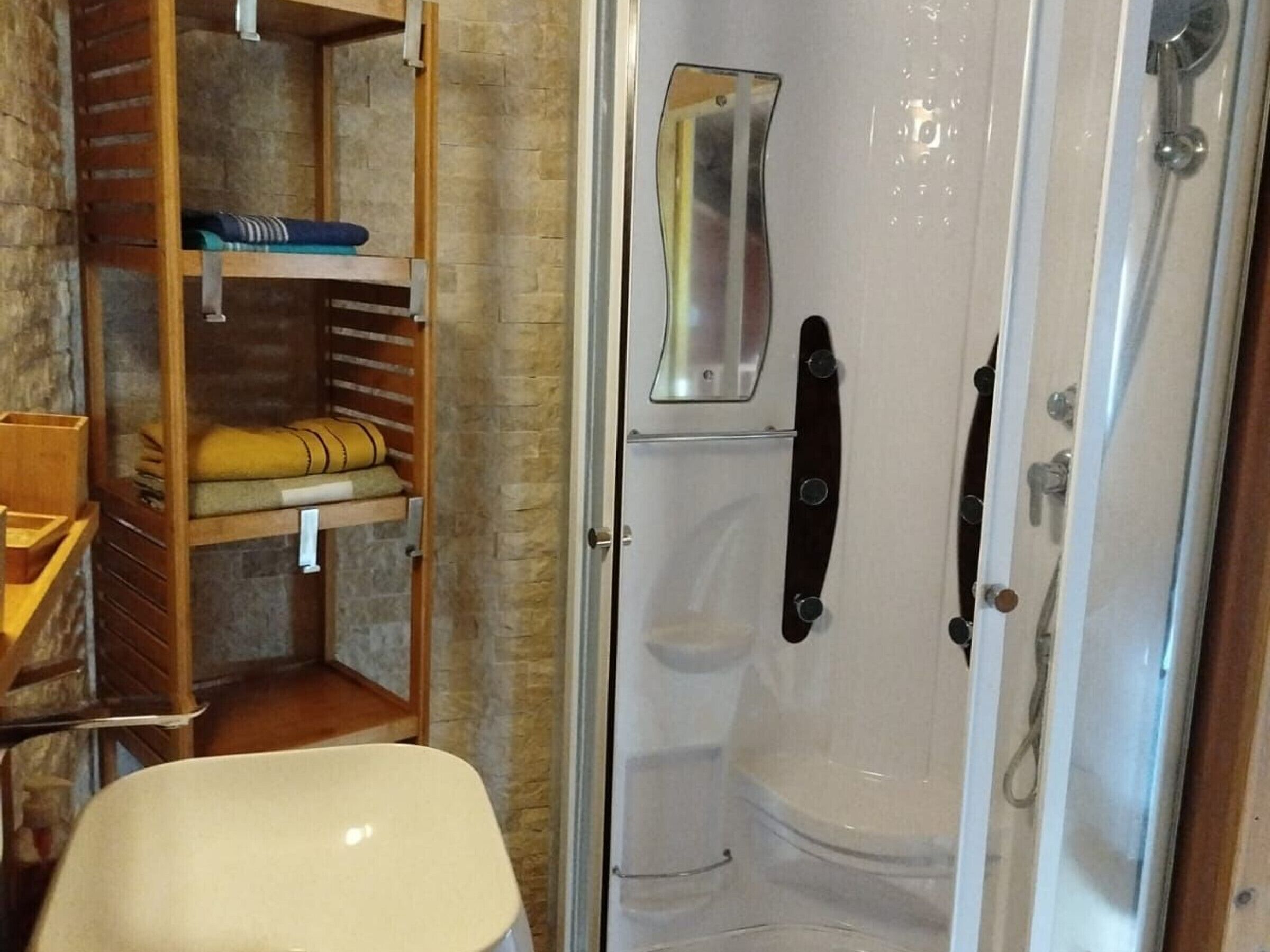 A bathroom with a shower, a wooden shelf with towels, and a toilet.