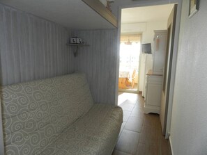 Et værelse med en stribet sofa, en lille reol med genstande og en døråbning til et køkkenområde.