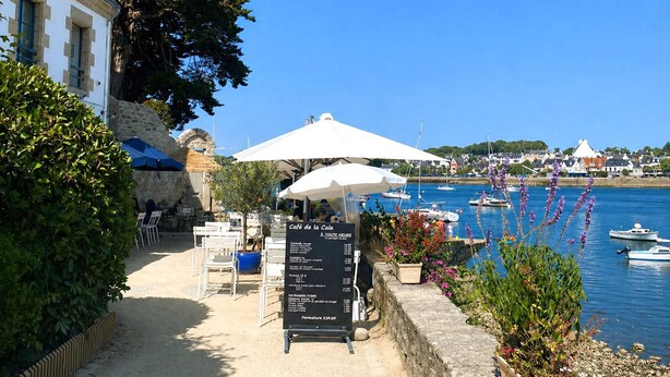 Un café en bord de mer avec une terrasse, un tableau affichant le menu et des bateaux en arrière-plan.