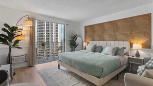 Ein modernes Schlafzimmer mit einem großen Bett, einem Holz-Kopfschild, einem Balkon mit Blick auf Hochhäuser und einer großen Küstengrün-Pflanze in einem Topf.