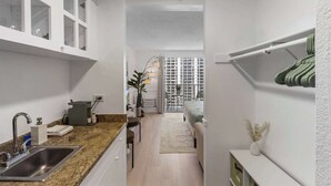 Eine schmale Küche mit Spüle, Schränken und einer Arbeitsplatte. Ein Essbereich mit Tisch und Stühlen lädt zum Verweilen ein, und durch ein Fenster ist ein Gebäude sichtbar.