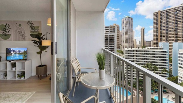 Ein Balkon mit Blick auf hohe Gebäude, ein kleiner Tisch mit Stühlen, ein Blumentopf und ein Fernseher auf einem weißen Schrank.