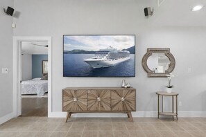 A room with a large framed picture of a cruise ship, a wooden cabinet, a round mirror, and a small table with a potted plant.