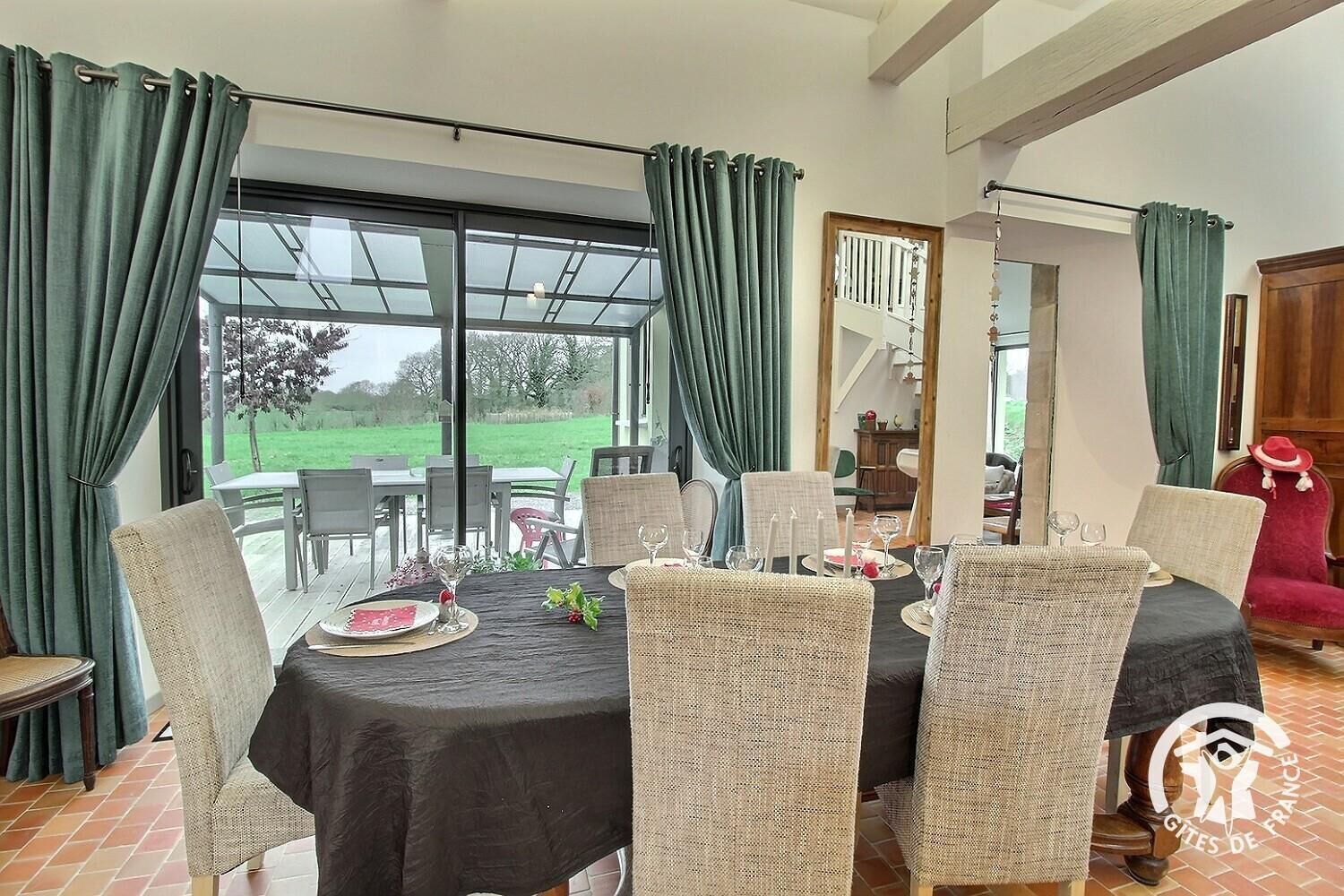Ein Essbereich mit einem Tisch, gedeckt für sechs Personen, umgeben von Korbstühlen, eine Glasveranda mit Blick auf eine grüne Landschaft und ein roter Sessel in der Ecke.