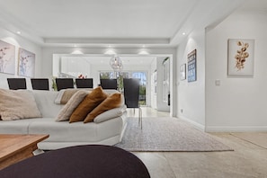 A modern living room with a white sofa, a coffee table, and a rug. There are framed pictures on the walls and a chandelier hanging from the ceiling.
