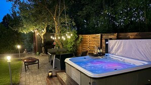 Ein Whirlpool mit einer Holzterrasse und Lichterketten, umgeben von einem Holzzaun und Bäumen.