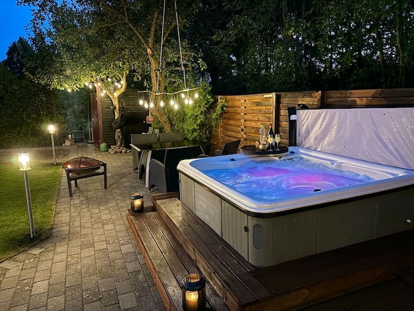 A hot tub with a wooden deck and string lights, surrounded by a wooden fence and trees.